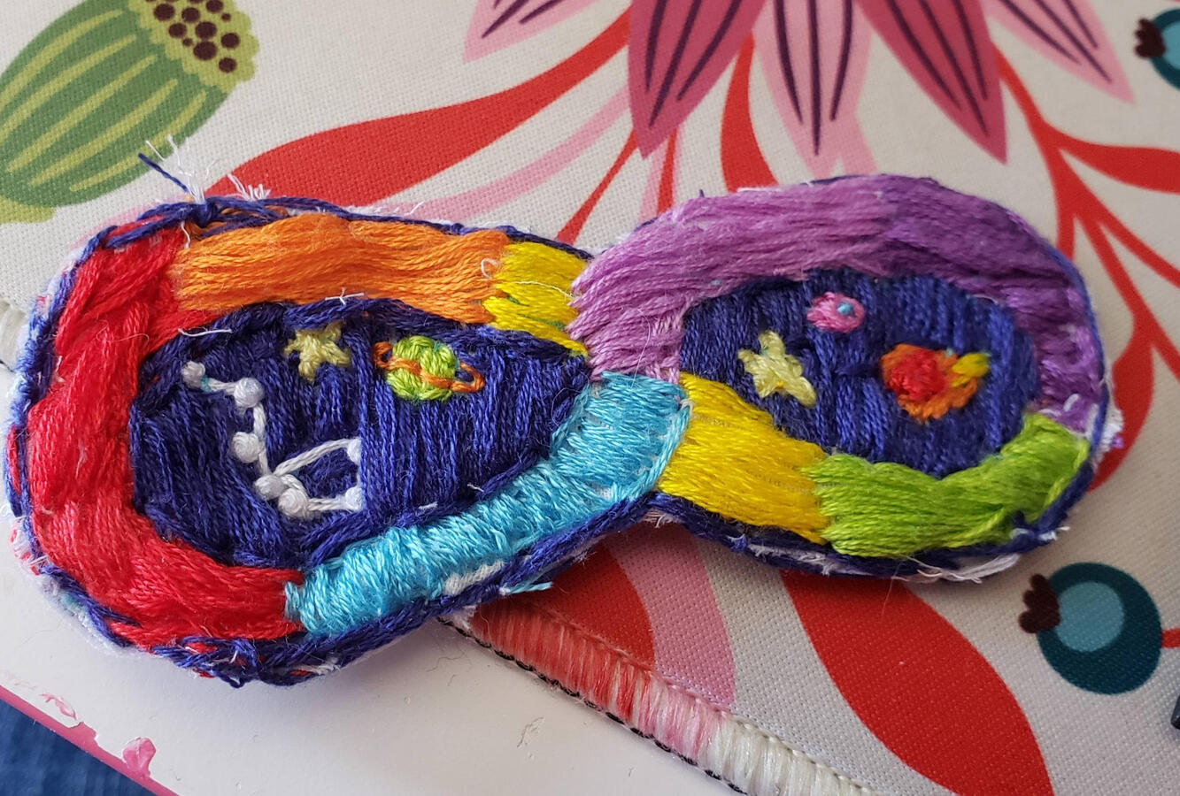 A hand embroidered patch of the neurodiversity loop. Inside the two negative spaces is a blue drop and handstitched stars, planets and comets. The big dipper is visible on one side.