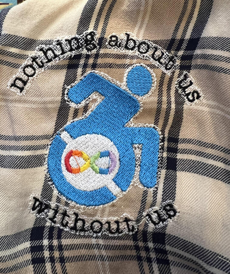 An embroidered shirt showing a blue wheelchair user shown mid push, with a neudiversity symbol in the wheel. the text surrounding reads "nothing about us without us"