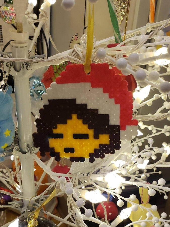 A perler bead ornament of "frisk" from undertale, notable nonbinary game protagonist. They wear a santa hat and have brown hair.